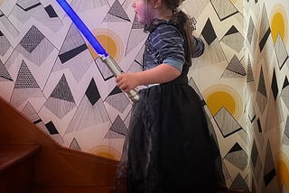 Small girl making her way upstairs wearing a black Halloween ballgown and holding a light saber.