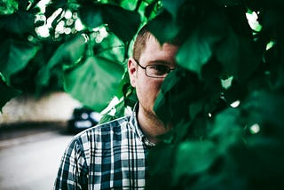 A man with eyeglasses standing behind green leaves.