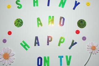A poster with a white background and the words “Shiny and Happy on TV”, decorated with smiley faces and daisy flowers