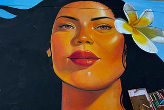 Chalk drawing of a woman’s face. The woman has long black hair, bright red lipstick, light brown skin, and a white flower in her hair. A box of pastel chalks is in the lower right corner of the photo.