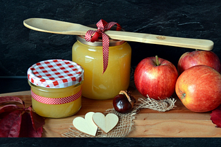 The makings of applesauce are arranged neatly on a wooden cutting board, including reddish-yellow apples, jars of chunky yellow sauce, a wooden spoon, with fall decor details like gingham ribbons around the spoon and one jar and as a pattern on the jar’s lid, two little wooden heart cut-outs, and a scrap of burlap.