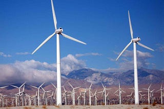 Two wind turbines shown close up, in front of a large wind farm with clouds in the background