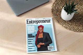 Magazine ‘Entrepreneur’ on a wooden table next to a closed laptop and small cactus plant.