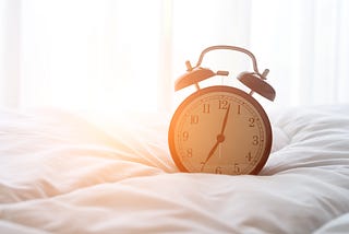 A clock sits on top of a white comforter with sun streaming in from the background