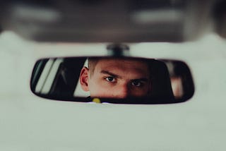 A young human with an intense look in their eyes looks at themselves in a car rearview mirror.