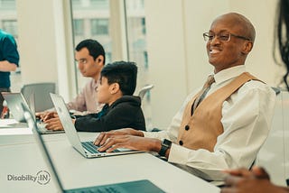 Smiling man with a learning disability works on a laptop in a meeting with other colleagues.