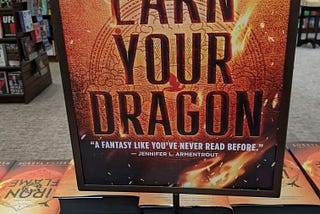A table in a bookstore showing a display of “Fourth Wing” and “Iron Flame” hardbacks. A printed sign placed in the center of the display reads “EARN YOUR DRAGON” in huge letters on an illustrated background with golden colors and flames.