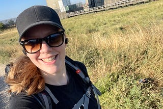 Me in a baseball cap, sunglasses, and an all-black outfit with a huge backpack, behind me the Dartford Creek Barrier.