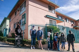 9 tenants stand in front of their building. One tenant holds a sign that says “WE ARE HERE TO STAY”