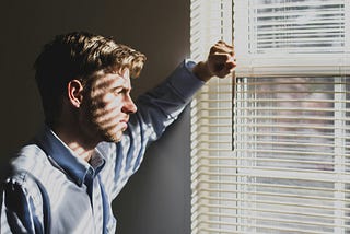 Solemn man looks out window with the blinds casting shadows on his face