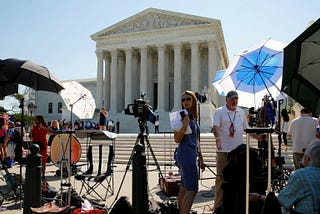 Photograph of the US Supreme Court, foregrounded by media cameras and reporters. The author is standing at the top of the steps in front of the SCOTUS building in a blue dress.