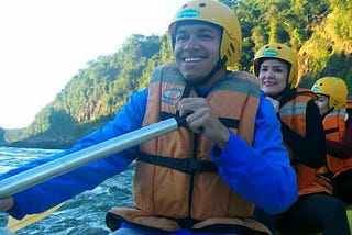 A man and two women paddle a kayak on a river. All three have big smiles and are  wearing yellow helmets and orange life vests. Tall green trees line the river on the right side of the photo.