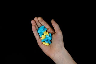 A person’s hand against a black backdrop, holding a mixture of blue and yellow pills.