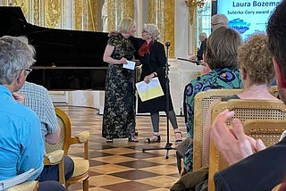 Two white women standing in front of a seated audience, one presenting an award to the other. A grand piano is visible in the background