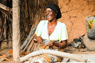 An elderly African woman sits outside a traditional mud-walled home, joyfully smiling and weaving natural fiber rope by hand. She wears a black headscarf, a light green blouse, patterned wrap skirt, and several bracelets. The setting is earthy and rustic, with palm fronds, a tree trunk, and household items around her, reflecting a rural environment and traditional craftsmanship.