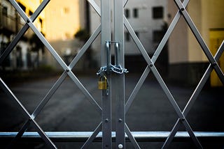 A metal gate kept locked by a padlock through which we can see a world we cannot reach.