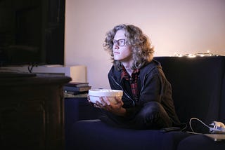 Person with light curly hair holding a popcorn bowl and illuminated by a TV screen they’re staring intently into