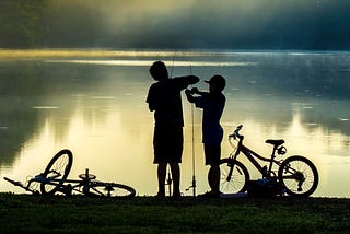 The silhouettes of two boys in front of a lagoon with their bicycles lying at their feet