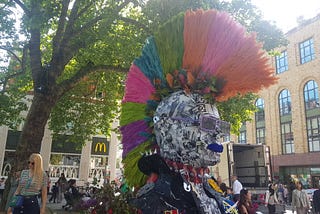 A head and shoulders of a punk with a rainbow-coloured mohawk surrounded by flowers and a bench set in a town square with trees and shops in the background and people milling around.