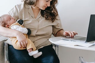 Woman holding baby in one arm while seated, looking at her laptop screen