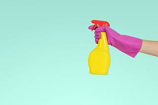 An arm in a pink plastic glove reaches into frame from the right side, holding a yellow spray bottle with a red top. The background is a light seafoam or turquoise.