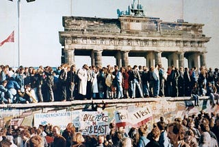 Color photograph of many people standing at the Bradenburg Gate at the Berlin Wall while a crowd below holds signs and banners.