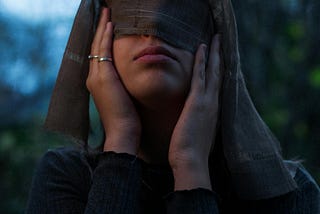 Image of a woman blindfolding her eyes with a brown scarf. She’s holding the scarf and her face.
