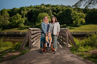 Genetic counselor, Leslie Walsh Cyprych, with Tracy Fisher and her daughter, Brie, smiling outside in front of a bridge