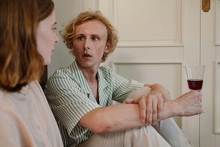 Man looking at a girl and holding a glass of wine — with a face that looks like he’s been asked an awkward question