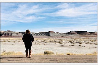 A plus size woman with midlength brown hair is standing on an empty road, dressed in all black and brown knee boots, facing away from the camera, toward a barren desert landscape with gray and red hills in the distance against a bright blue sky.