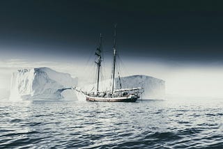 A small sailing ship passes a towering iceberg across calm polar waters beneath a dark, expansive sky, evoking uncertainty, exploration, and navigation.