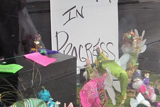 A street scene reflected in a store window highlights a “work in progress” sign in Boots Davidovitch’s story on creativity, AI, and Linklater’s two newest films, Nouvelle Vague and Blue Moon.