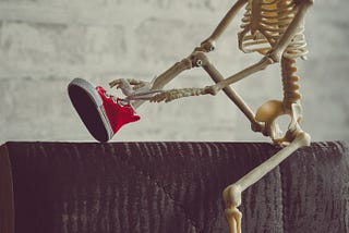 An adorable skeleton sits on a ledge with a red cap on, tying their red high-top converse sneakers