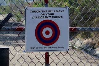 Sign on a fence with a bulls-eye that says “touch the bulls-eye or your lap doesn’t count.”