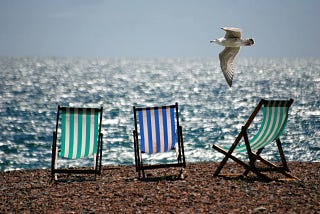 Three-green and blue stripy beach chairs on a pebbled beach with glittering waves in the background and a seagull flying by.