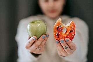 A blurred, out of focus person holding up an apple on the right and a glazed donut with a few sprinkles with a bite taken out of it on the left.