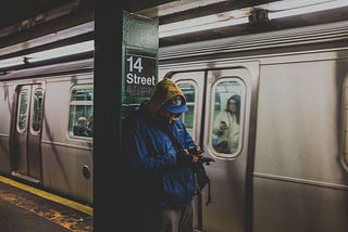 Man in blue hoodie using a smartphone in 14 Street station with a train nearby.