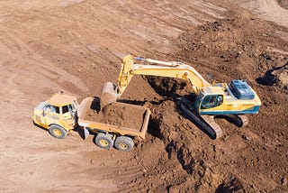 An articulated dump truck is to the left in the photo and positioned lower than the hydraulic excavator which is loading it from the right and above. The excavator is emptying a load of dirt from its bucket into the truck, which already is holding some dirt.