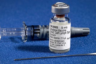 Close-up photo of three components of a smallpox vaccination kit with the vial of vaccine in the foreground on a blue background
