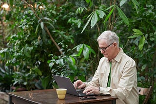 Elderly man sitting at a table with a laptop typing. There is a yellow cup behind the screen and green foliage around him.