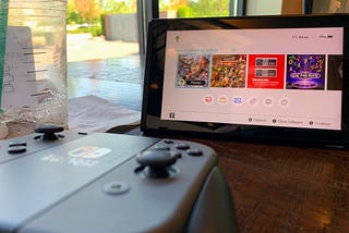 A Nintendo Switch propped up in “kickstand mode” on a table in a coffee shop, with a mostly empty cup and a controller sitting nearby. A window is visible in the background with some sun, trees, and a parking lot in view.