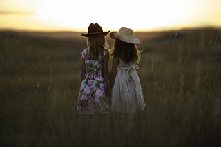 Sibling girls in summer dresses standing in a field facing either sun rise or sunset.