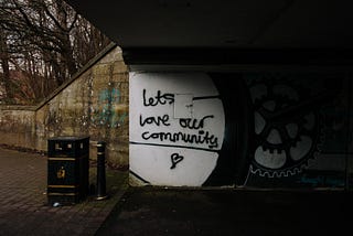 graffiti in black paint on white tunnel wall, “Lets love our community” with a heart below.