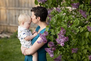 mother kissing boys forehead, standing near purple lilacs