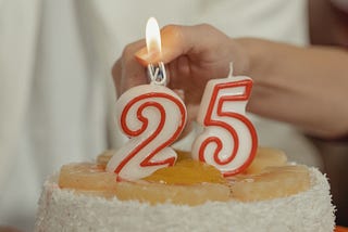 White Numbered birthday candles lined with red for the number twenty five sits atop a white coconut cake with a caramel top. The number two is being lit with a lighter.