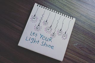 Picture of a notebook on a wooden table. There are drawn lightbulbs on the notebook and the words Let Your Light Shine.