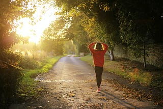 A female runner walking with hands on her head facing the rising sun on a leafy road