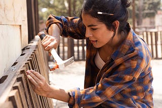Young woman wearing a loose checked shirt, pencil stuck in her hair, hammering a nail into a bedframe.
