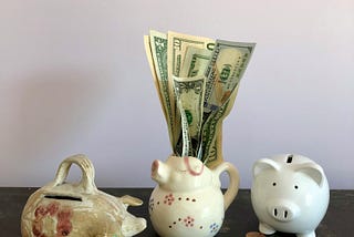 Three piggy banks on a table, with piles of coins and bills stuck in one bank.