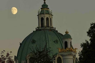 A grand domed building with classical architecture, featuring a green copper dome topped by a cupola and cross, stands partially obscured by trees. Ornate golden embellishments and statues accent the structure, especially on a turret to the right. A nearly full moon glows in the muted gray sky, suggesting dusk or dawn, adding a serene contrast to the building’s opulence.